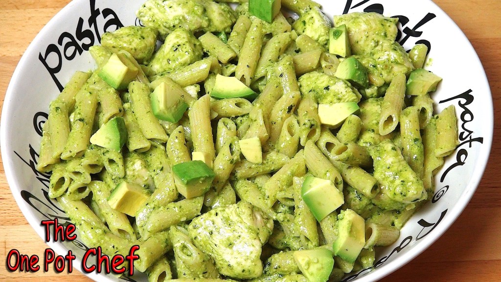 A bowl of green pesto pasta with pieces of chicken and avocado, served on a decorative plate with 'pasta' written around the rim.