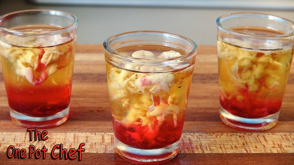 Three clear glasses filled with a layered drink featuring a red base and golden top, with floating edible flower petals.