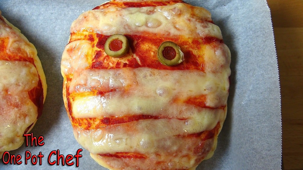 A baked pizza shaped like a mummy, featuring cheese and tomato sauce with olive eyes, displayed on parchment paper.