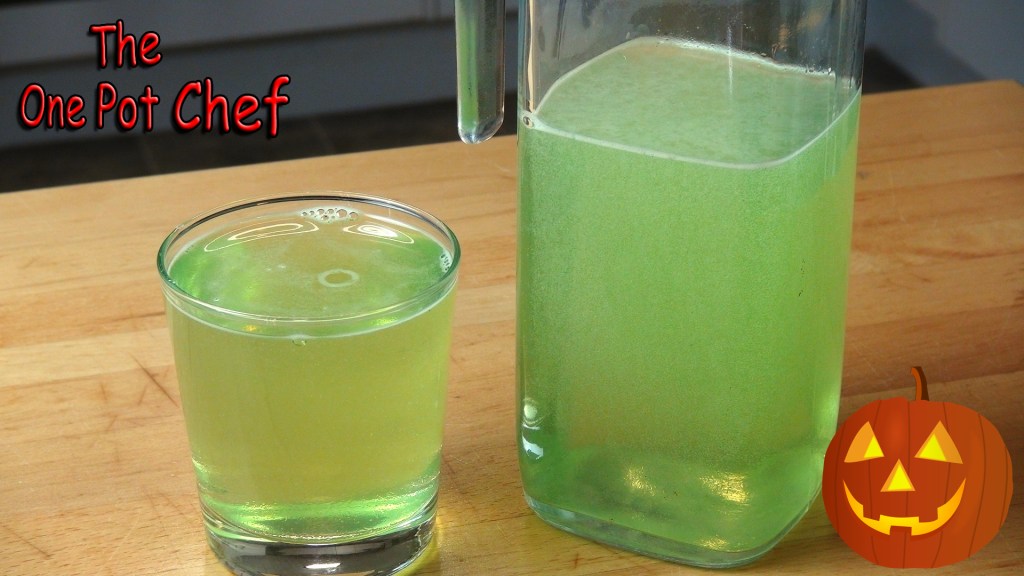 A glass and a pitcher containing a green fizzy drink on a wooden surface, with a Halloween pumpkin decoration in the corner.