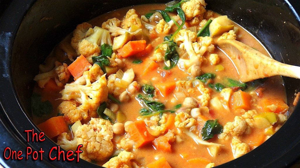 A pot of vegetable stew containing cauliflower, carrots, and spinach, with a creamy sauce and a wooden spoon resting in it.